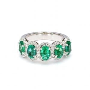 Front view of ring. A row of five oval cut emeralds are set along the front half of a simple white gold band with pave diamond halos around each.