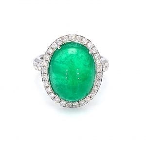 Front view of ring. A center cabochon emerald is haloed by round brilliant cut diamonds, with additional round brilliant cuts diamonds accenting half way down each side of a white gold shank.