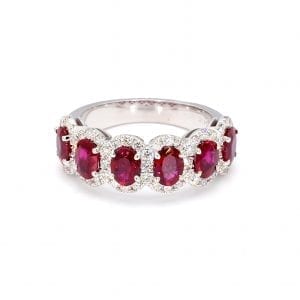 Front view of ring. A row of six oval cut rubies band across the front half of a simple white gold band with interlocking pave diamond halos.