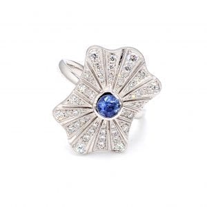 Front view of ring. A burst style mounting has a center light blue sapphire with rows of graduated diamonds fanning out from the middle. Attached to a simple white gold band.