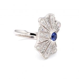 45 degree angle of ring. A burst style mounting has a center light blue sapphire with rows of graduated diamonds fanning out from the middle. Attached to a simple white gold band.