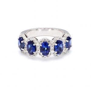 Front view of ring. A row of five oval cut, blue sapphires are set along the front half of a simple white gold band with interlocking pave diamond halos.