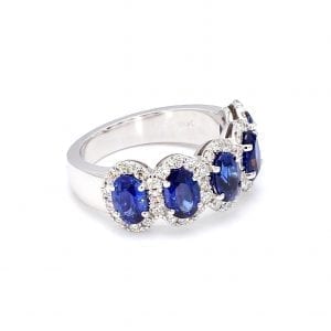 45 degree angle of ring. A row of five oval cut, blue sapphires are set along the front half of a simple white gold band with interlocking pave diamond halos.