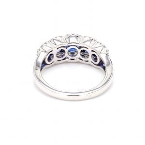 Back view of ring. A simple white gold band leads to a setting that holds five round sapphires framed by pave diamond scalloped edges.