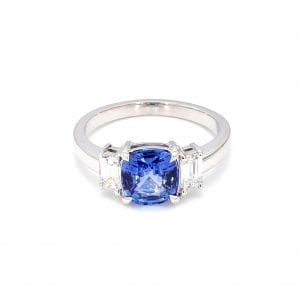 Front view of ring. A center cushion-cut sapphire is accented with an emerald cut diamond on either side, attached to a simple white gold band.