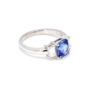 45 degree angle of ring. A center cushion-cut sapphire is accented with an emerald cut diamond on either side, attached to a simple white gold band.