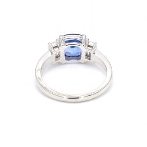 Back view of ring. A simple white gold band leads to the setting for a three stone ring with a cushion cut sapphire center and an emerald cut diamond on either side.