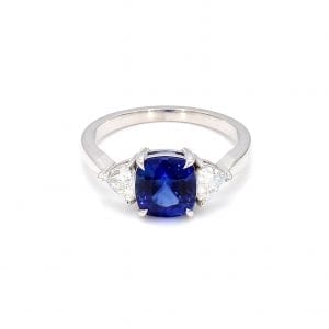 Front view of ring. A three stone ring showcases a center cushion cut sapphire with a trillion cut diamond on either side, attached to a simple white gold band.