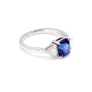 45 degree angle of ring. A three stone ring showcases a center cushion cut sapphire with a trillion cut diamond on either side, attached to a simple white gold band.