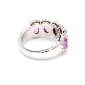 135 degree view of ring. A simple white gold band leads to a five stone oval setting that has five pink sapphires with pave diamond halos.