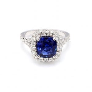Front view of ring. A center cushion cut sapphire is surrounded by a halo of pave diamonds with additional diamonds set halfway down a split shank.