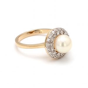 A ring is shown at a 45 degree angle. A center round, white pearl center is encircled by a pave diamond halo, attached to a simple yellow gold band.