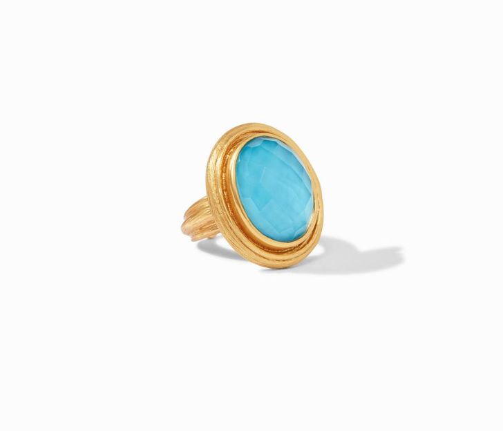 45 degree angle of ring. An oval, domed pacific blue stone center is haloed by a textured frame of gold plating, Attached to a matching textured and plated shank.