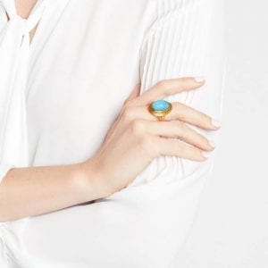 An oval, domed pacific blue stone center is haloed by a textured frame of gold plating, Attached to a matching textured and plated shank. the ring is pictured on the middle finger of a lady with her arms crossed, showing off her hand.