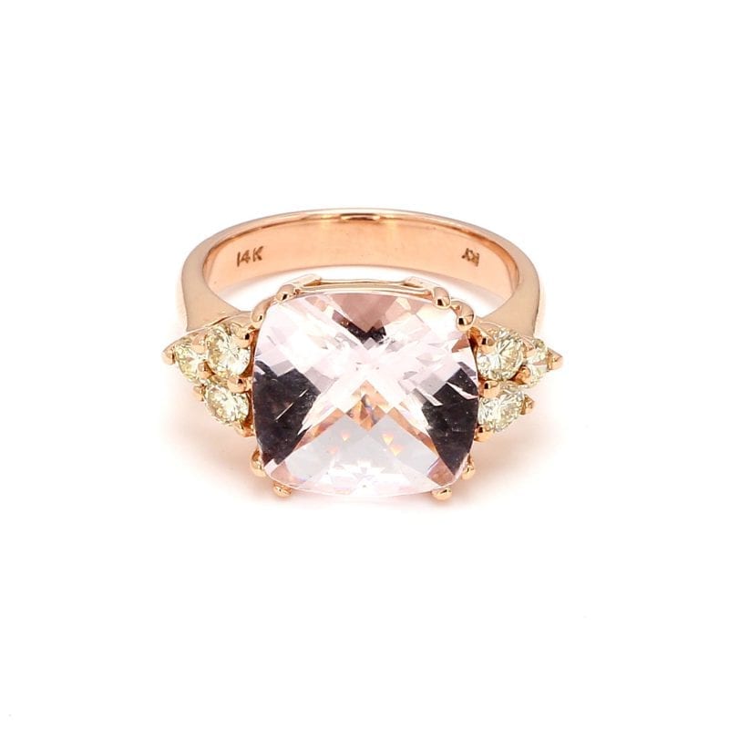 Front view of ring. A pale pink, cushion cut morganite sits in the center with a trio of round brilliant cut diamonds forming a side triangle motif on each side. This ring is complimented by a simple rose gold shank.