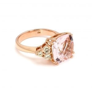 45 degree angle of ring. A pale pink, cushion cut morganite sits in the center with a trio of round brilliant cut diamonds forming a side triangle motif on each side. This ring is complimented by a simple rose gold shank.