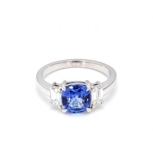 Front view of ring. A center cushion-cut sapphire is accented with an emerald cut diamond on either side, attached to a simple white gold band.