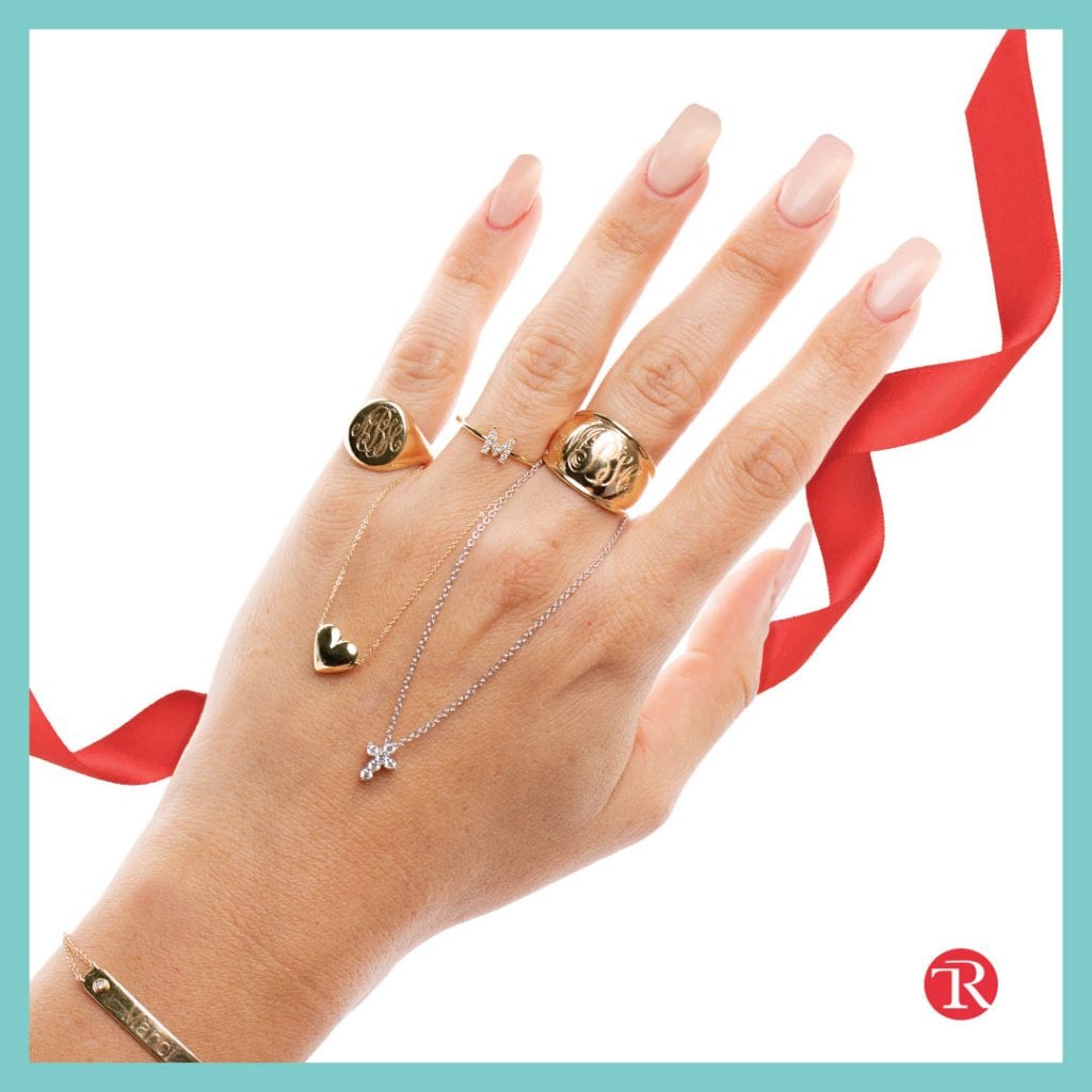 Picture of hand with ribbon in the background. Hand is modeling the express yourself jewelry trend jewelry. Hand has a ring on three fingers and a bracelet on the wrist. A heart necklace hangs from the ring finger and a cross necklace hangs from the middle finger.