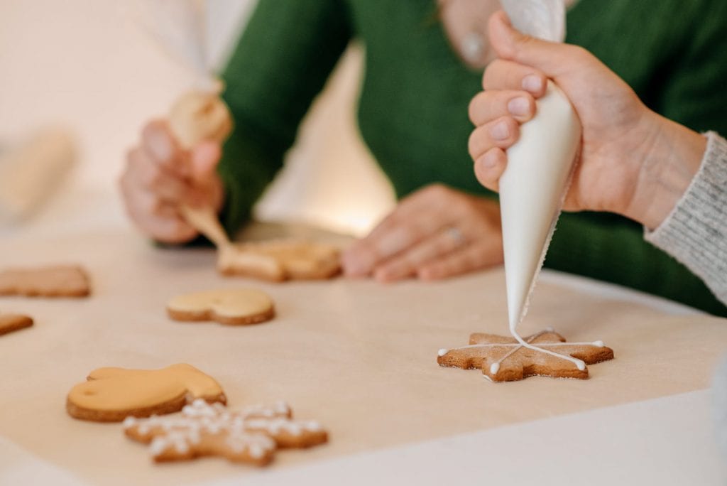 Two people decorating christmas cookies. Just the peroples hands are shown. Hands are holding icing bags. 
