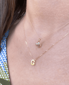 Two gold necklaces on a woman's chest. The top neckle is a love know. The bottom necklace is a letter "G" pendant.