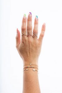 Hand with different colors on the nails. Hand is weraing three "x" shaped rings. Three gold bracelets on the arm. One bracelet features a row of rainbow gems.