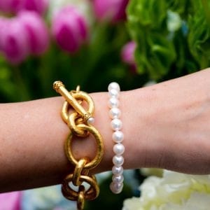 woman's wrist in front of flowers with gold link bracelet and pearl bracelet